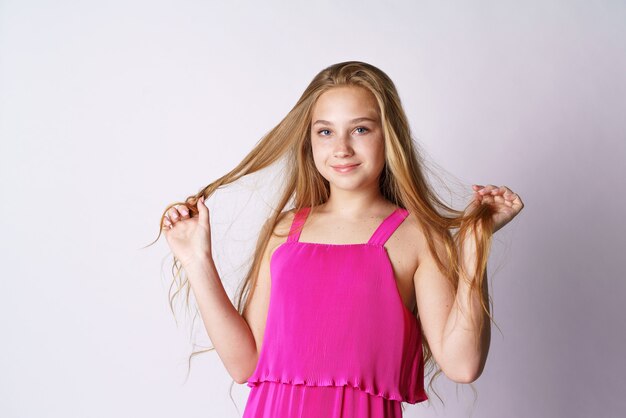 A cute and happy girl of caucasian appearance in pink clothes on a white wall plays with her long hair