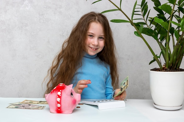 테이블에 있는 귀여운 행복한 소녀는 계산기 금융 문해력에 대한 돼지 저금통의 돈을 계산합니다