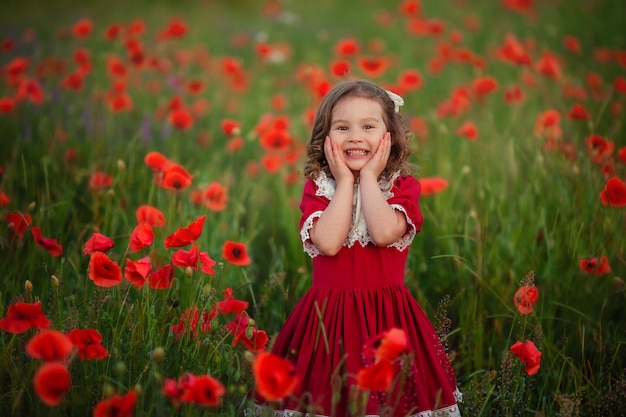 Ragazza felice sveglia con capelli ricci in un vestito d'annata rosso con pizzo nei raggi del tramonto su un campo del papavero.