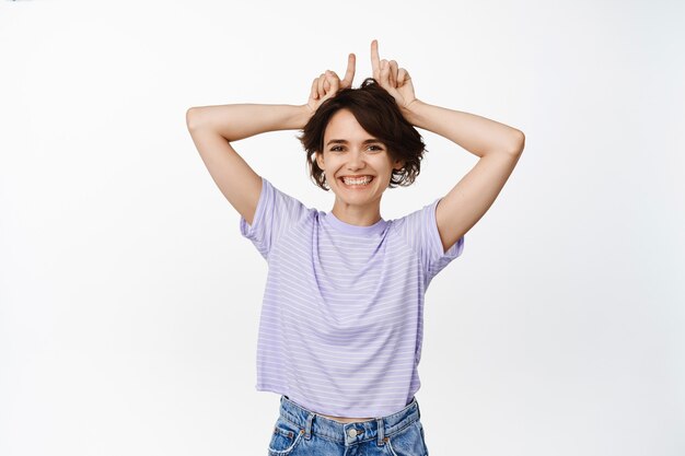 Cute happy girl, young woman smiling, showing bull devil horns, fingers on head and laughing, standing silly on white