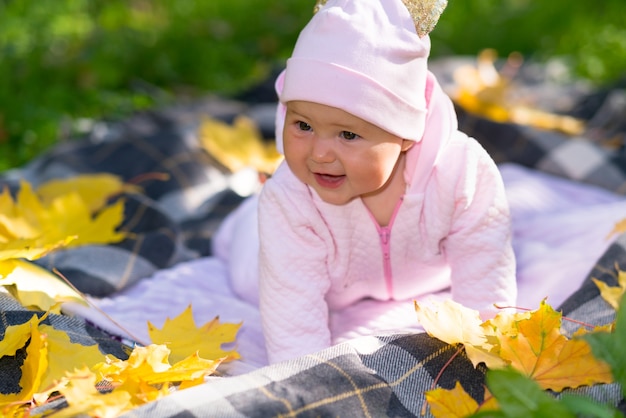 Cute happy little baby girl in a pink outfit playing outdoors in autumn on a rug on the grass surrounded by colorful yellow leaves