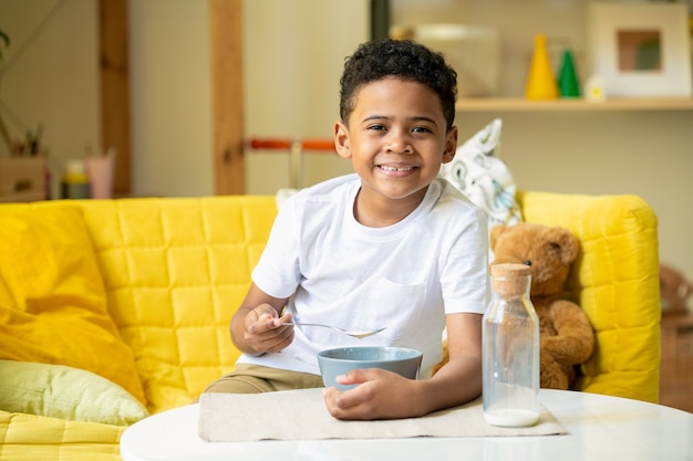 Cute happy little boy of African ethnicity with toothy smile while sitting on couch and eating cornflakes with milk for breakfast