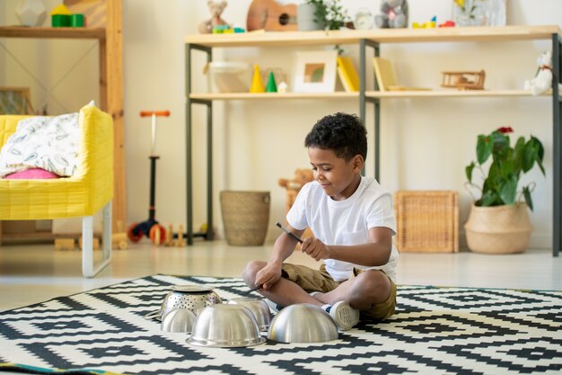 Cute happy little boy of elementary age sitting on the floor of living-room and beating on tops of metallic bowls in home environment