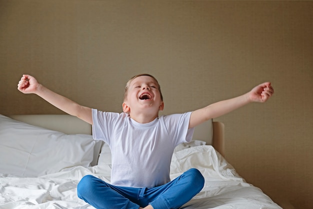 cute happy little boy stretching and yawning on white bed in a bedroom