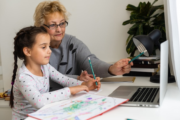 Cute and happy little girl child using laptop computer with her grandma, studying through online e-learning system.