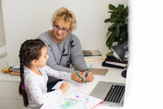 Cute and happy little girl child using laptop computer with her grandma, studying through online e-learning system.