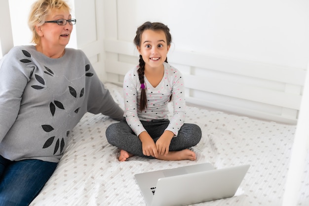 Cute and happy little girl child using laptop computer with her grandma, studying through online e-learning system.