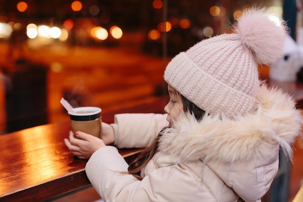 Cute happy little girl drinks hot tea in a street cafe