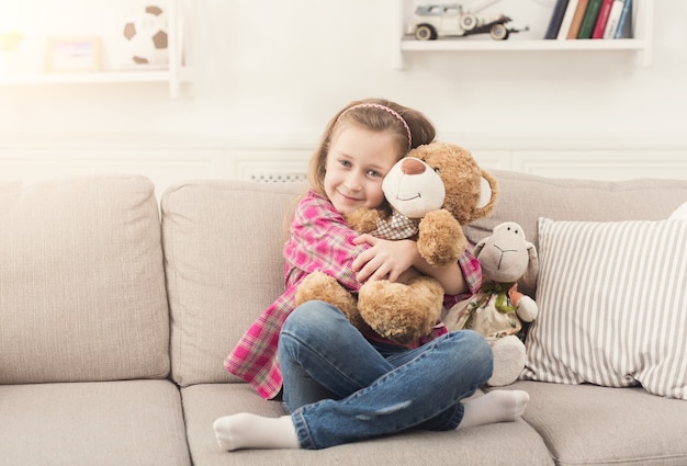 Cute happy little girl embracing teddy bear. Pretty female kid at home, sitting on sofa with her favorite toy, copy space