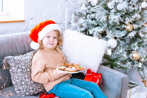 Cute happy little girl with curls is sitting on chair near christmas tree with plate of christmas