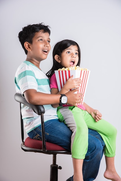 Cute happy little Indian boy and girl or siblings eating popcorn and watching television while sitting on chair