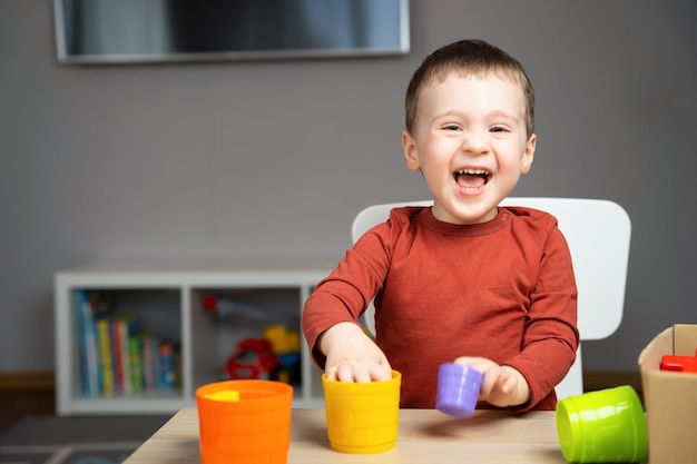 2 歳のかわいい幸せな小さな幼児の男の子が子供たちのテーブルに座って、自分の部屋で色とりどりのピラミッドで遊んでいます子供用の教育玩具ソフト選択フォーカス