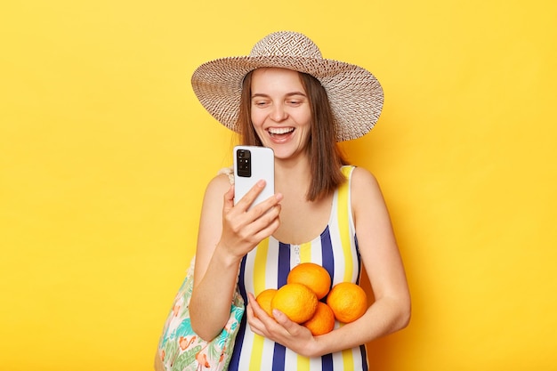 Cute happy positive woman wearing striped swimsuit and straw hat isolated yellow background standing with fruit using mobile phone chatting with friends from resort