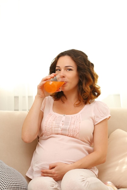 Cute happy pregnant woman on cozy sofa with glass of juice in the room close up