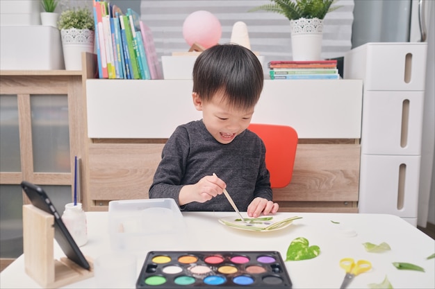 Il piccolo ragazzo asiatico sorridente felice sveglio del bambino gode di usando la colla che fa le arti a casa