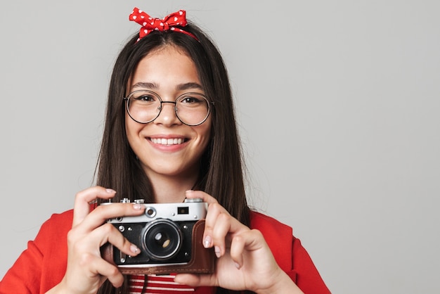 Милая счастливая девочка-подросток в повседневной одежде стоит изолированно над серой стеной и фотографирует с помощью портретной камеры