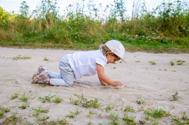 cute happy toddler in a cap crawls on all fours along a country road in the sand on a sunny summer day.