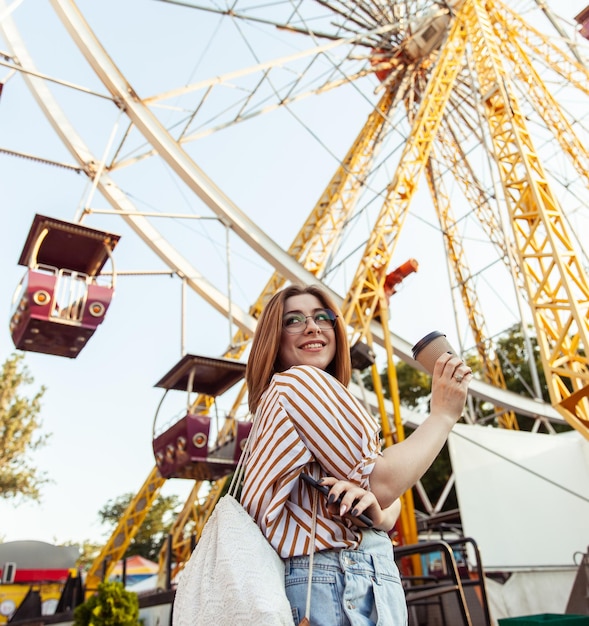 Foto bella donna felice con occhiali e tuta di denim che tiene una tazza di caffè in un parco divertimenti