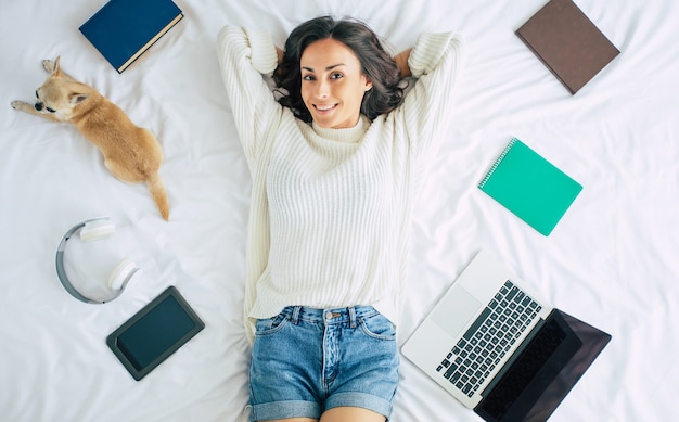 Cute Happy young beautiful woman is lying on bed at home with her little dog and some stuff for study. Girl with laptop and phone