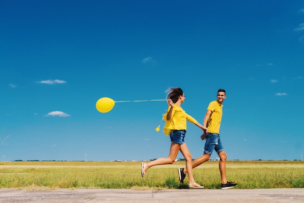 Giovani coppie felici sveglie che si tengono per mano e che corrono nel campo mentre la ragazza sta tenendo il pallone giallo in sua mano.