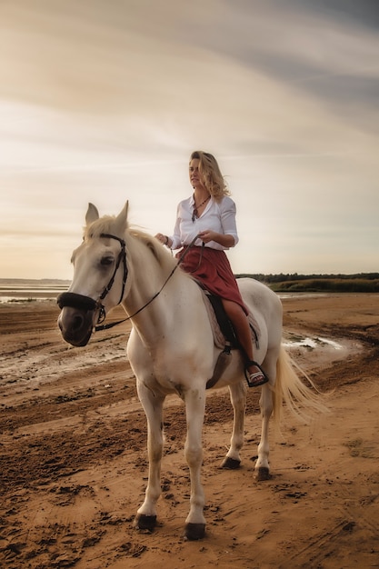 Cute happy young woman on horseback in summer beach by sea. Rider female drives her horse in nature on evening sunset light background. Concept of outdoor riding, sports and recreation. Copy space