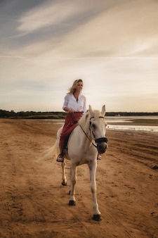 Carina giovane donna felice a cavallo in estate spiaggia dal mare. la femmina del cavaliere guida il suo cavallo in natura sullo sfondo della luce del tramonto serale. concetto di equitazione all'aperto, sport e tempo libero. copia spazio