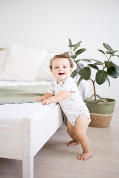 A cute healthy baby boy in a spacious bright bedroom is standing next to a bed with white linens the child is standing with support the child is learning to walk