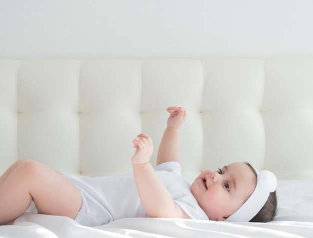 Cute healthy baby girl 6 months smiling in a white bodysuit lying on a bed on white bedding.