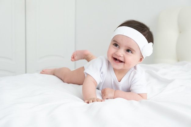 Cute healthy baby girl 6 months smiling in a white bodysuit lying on a bed on white bedding.