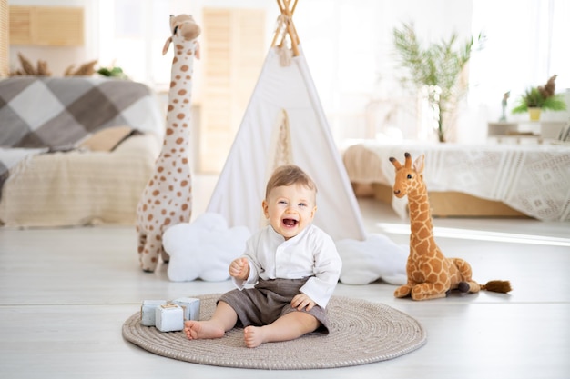 A cute healthy boy is sitting on a rug in the bright living room of the house against the background of a wigwam and plush toys playing with wooden educational toys home textiles