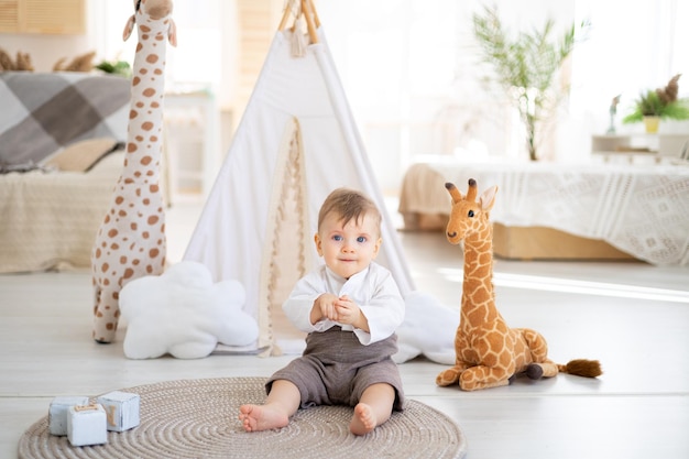 A cute healthy boy is sitting on a rug in the bright living room of the house against the background of a wigwam and plush toys playing with wooden educational toys home textiles