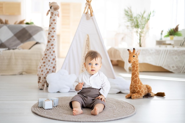 A cute healthy boy is sitting on a rug in the bright living room of the house against the background of a wigwam and plush toys playing with wooden educational toys home textiles