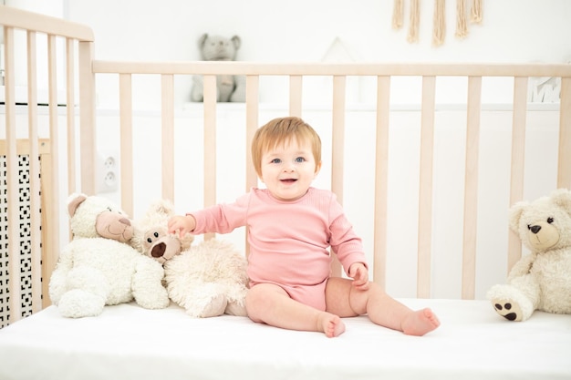 Cute healthy girl sitting on a baby cot with teddy bears on white bed linen in the bedroom at home baby sleeping in her crib