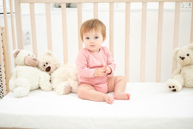 Cute healthy girl sitting on a baby cot with teddy bears on white bed linen in the bedroom at home baby sleeping in her crib