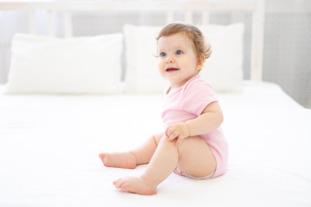 A cute healthy little girl in a pink bodysuit is sitting on the bed on white bed linen smiling