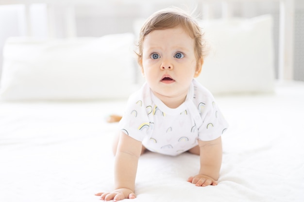 Cute healthy little girl in a white bodysuit crawls on the bed, on white bed linen