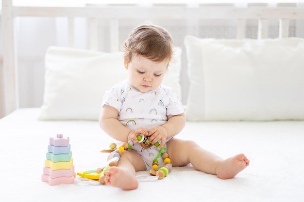 흰색 바디슈트를 입은 귀엽고 건강한 어린 소녀가 어린 아이들의 초기 발달을 위해 장난감을 가지고 놀며 웃고 있는 흰색 침대 린넨에 침대에 앉아 있습니다.