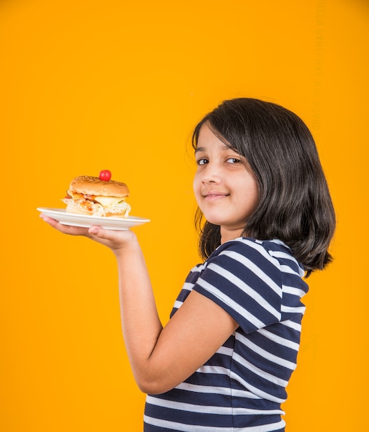 Милая индийская или азиатская маленькая девочка ест вкусный гамбургер, сэндвич или пиццу в тарелке или коробке. Стоя изолированные на синем или желтом фоне.
