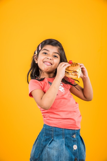 Cute indian or asian little girl eating tasty burger, sandwich or pizza in a plate or box. standing isolated over blue or yellow background.