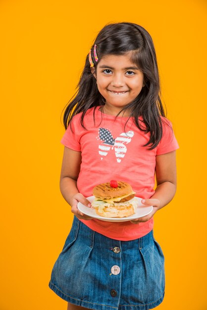 Cute Indian or Asian little girl eating tasty Burger, Sandwich or Pizza in a plate or box. Standing isolated over blue or yellow background.