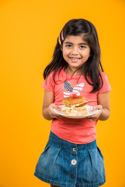 プレートまたはボックスでおいしいハンバーガー、サンドイッチまたはピザを食べるかわいいインドまたはアジアの少女。青または黄色の背景の上に孤立して立っています。