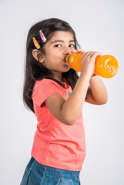 Cute Indian or Asian little girl with a pet bottle of Orange or Mango cold drink or fruit juice, drinking or holding while standing isolated over blue or white background.