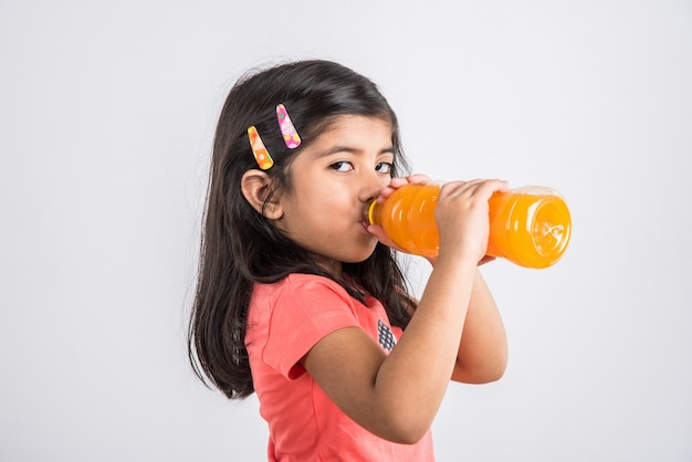 Cute Indian or Asian little girl with a pet bottle of Orange or Mango cold drink or fruit juice, drinking or holding while standing isolated over blue or white background.