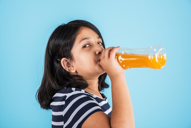 オレンジまたはマンゴーの冷たい飲み物またはフルーツジュースのペットボトルを持って、青または白の背景の上に孤立して立っている間飲んだり、保持したりするかわいいインドまたはアジアの少女。