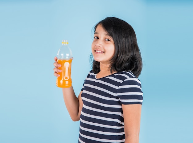 Cute indian or asian little girl with a pet bottle of orange or mango cold drink or fruit juice, drinking or holding while standing isolated over blue or white background