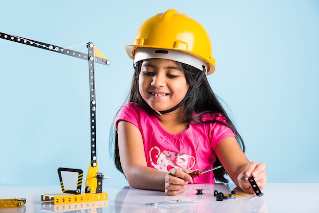 Cute indian baby girl playing with toy crane wearing yellow construction hat or hard hat, childhood and education concept