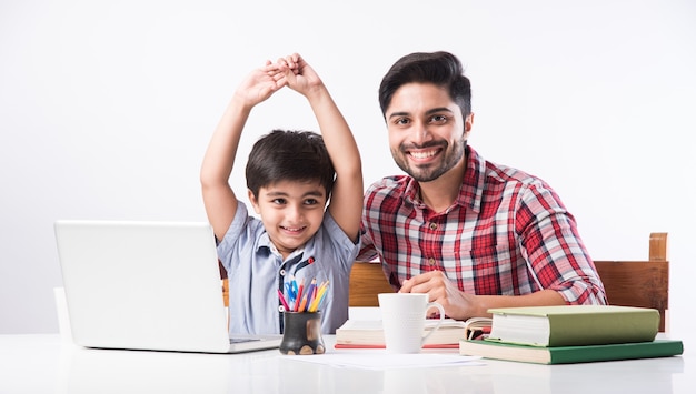 노트북과 책을 사용하여 집에서 숙제를하는 아버지 또는 남성 교사와 귀여운 인도 소년-온라인 교육 개념