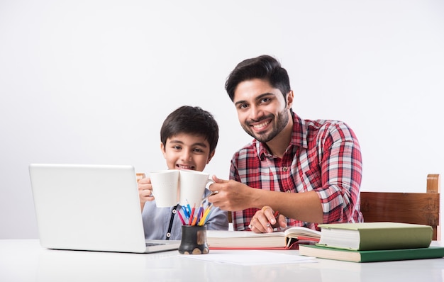 ノートパソコンと本を使用して自宅で宿題をしている父または男性の家庭教師とかわいいインドの少年-オンライン教育の概念