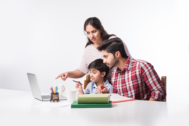 ノートパソコンと本を使用して自宅で勉強または宿題をしている父と母とかわいいインドの少年-オンライン教育の概念