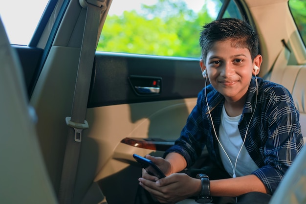 Cute indian child sitting in car and using smart phone and headphones gadget